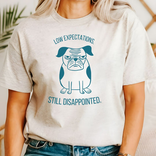 Gray t-shirt with a graphic of a dog and text 'Low expectations still disappointed' on a beige background.