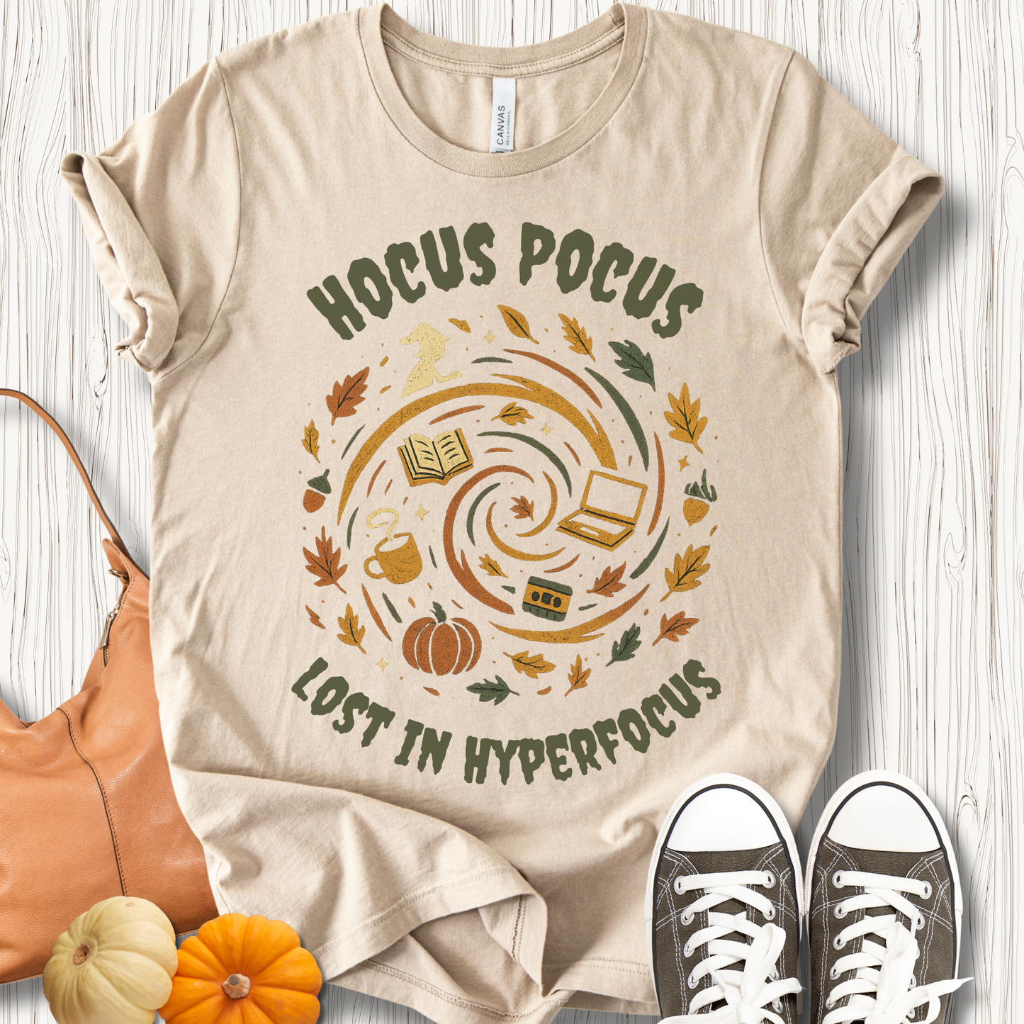 Beige t-shirt with 'Hocus Pocus Lost in Hyperfocus' design on a wooden surface with shoes and pumpkins.