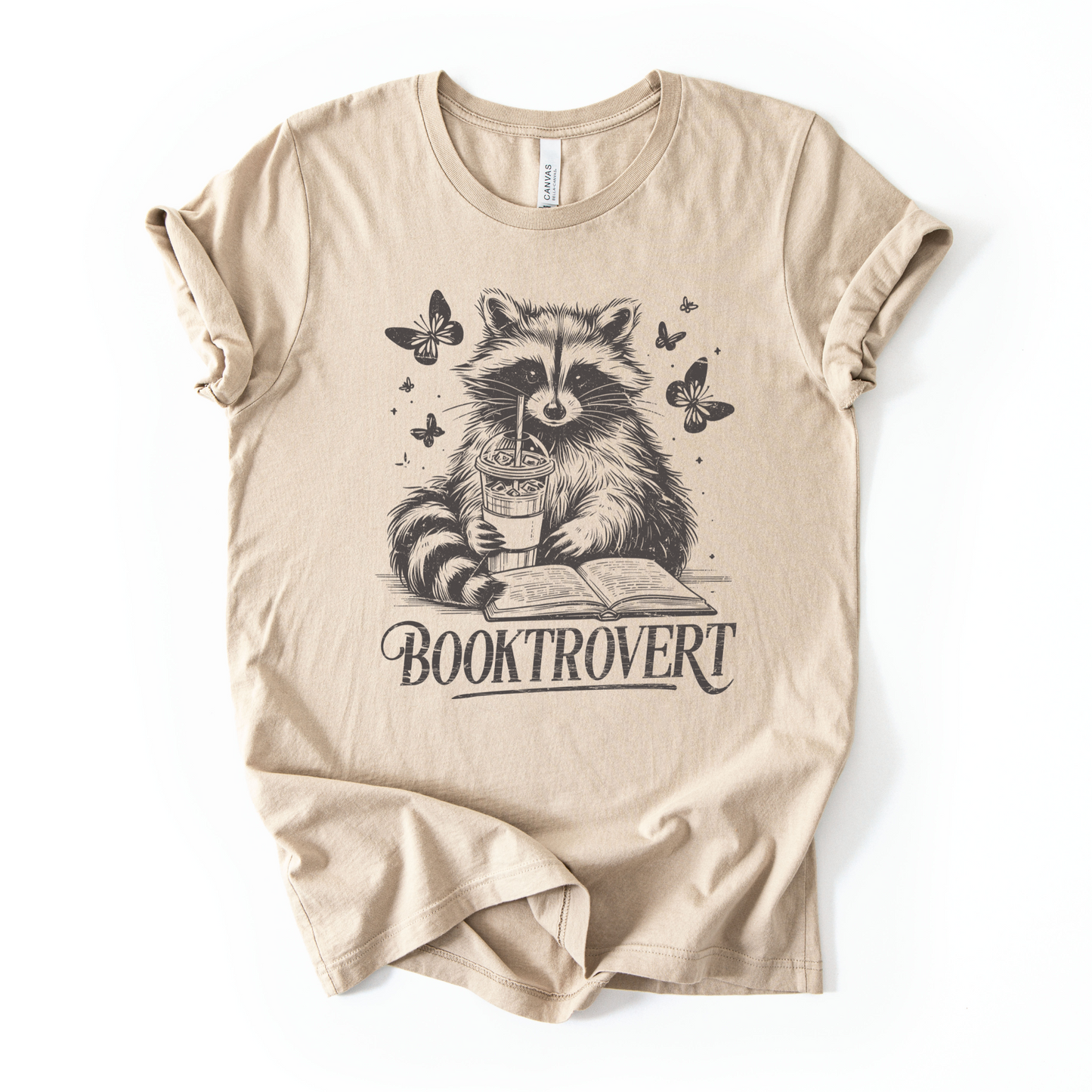 Beige t-shirt with a raccoon holding a book and cup design on a white background for book lovers with text that says "Booktrovert"