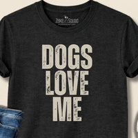 Black t-shirt with 'DOGS LOVE ME' text, jeans, and sneakers on a beige background