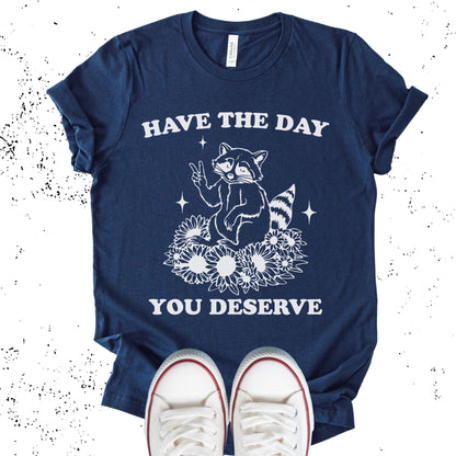 Unisex Zeke Squad T-shirt with the sarcastic phrase “Have the Day You Deserve” and a funny raccoon graphic.