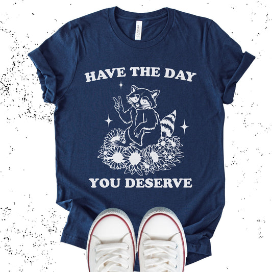 Unisex Zeke Squad T-shirt with the sarcastic phrase “Have the Day You Deserve” and a funny raccoon graphic.