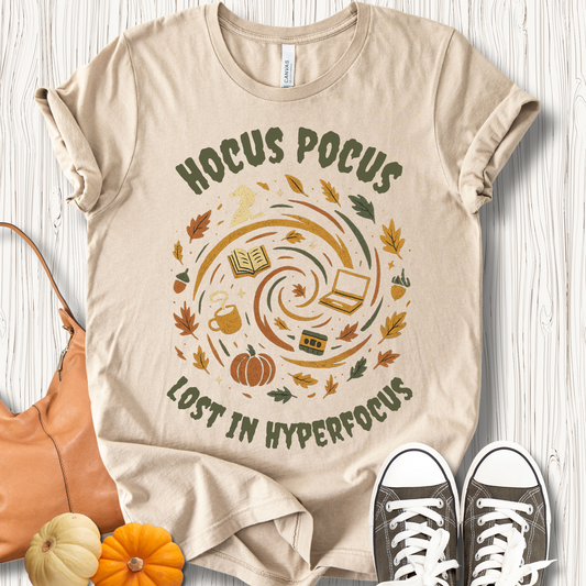 Beige t-shirt with 'Hocus Pocus Lost in Hyperfocus' design on a wooden surface with shoes and pumpkins.
