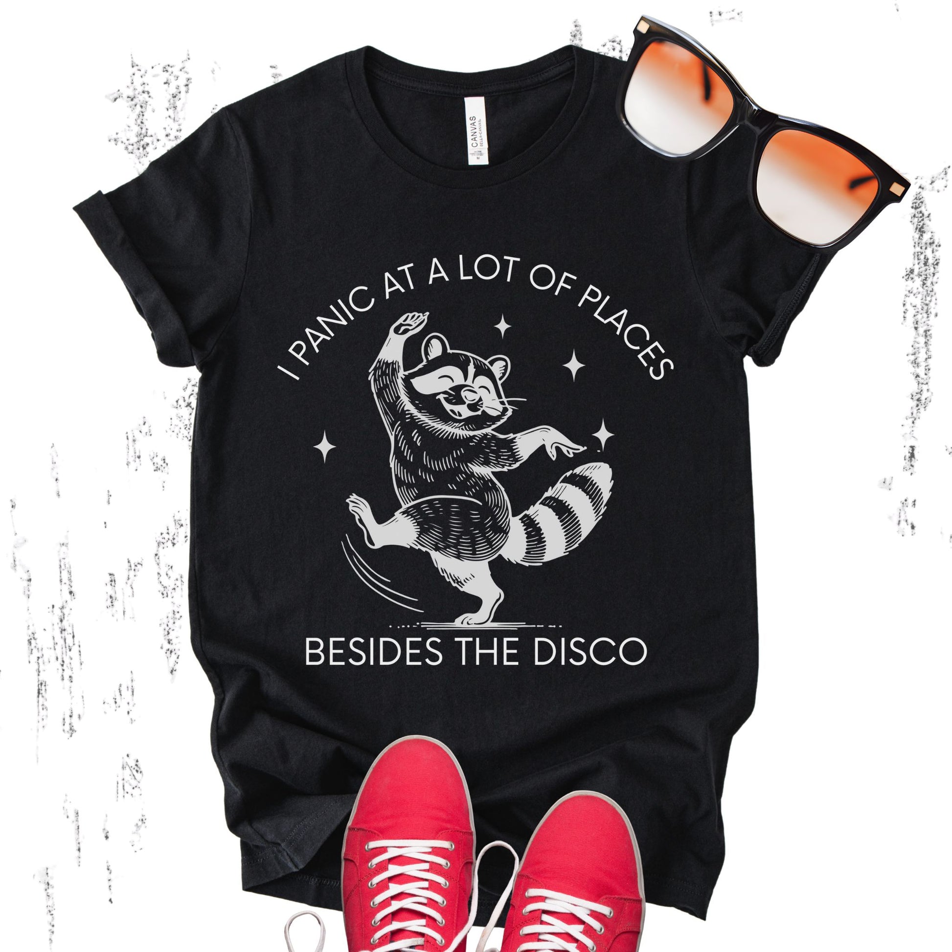 Unisex Zeke Squad T-shirt with the phrase “I panic at a lot of places, besides the disco” and a graphic of a dancing raccoon.