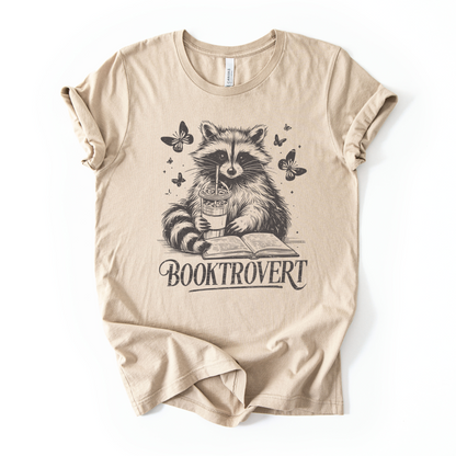 Beige t-shirt with a raccoon holding a book and cup design on a white background for book lovers with text that says "Booktrovert"
