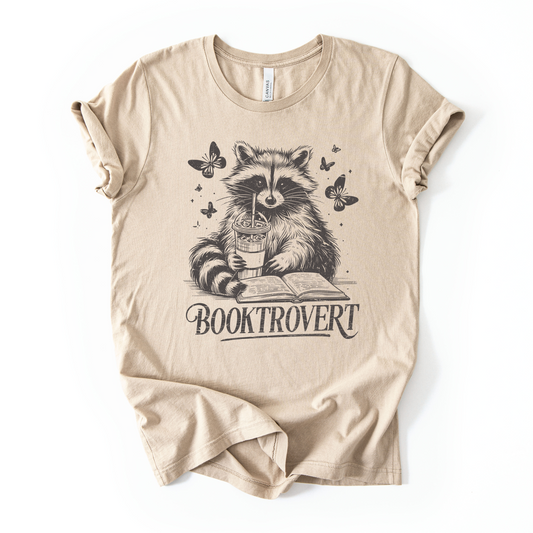 Beige t-shirt with a raccoon holding a book and cup design on a white background for book lovers with text that says "Booktrovert"